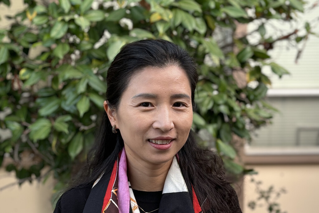 Estelle Zhao is a woman standing in front f a tree smiling with a multi-color silk scarf